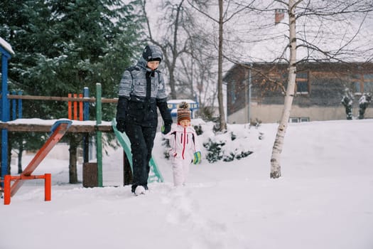 Mom and little daughter walk holding hands past a snowy slide under snowfall. High quality photo