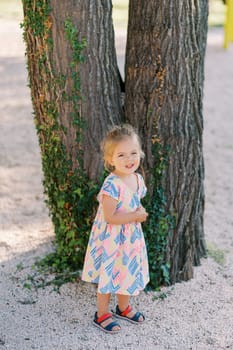 Little smiling girl stands near a tree covered with ivy. High quality photo