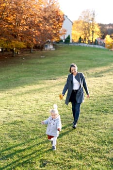 Mom with a bouquet of yellow leaves follows a running little girl through a green meadow. High quality photo
