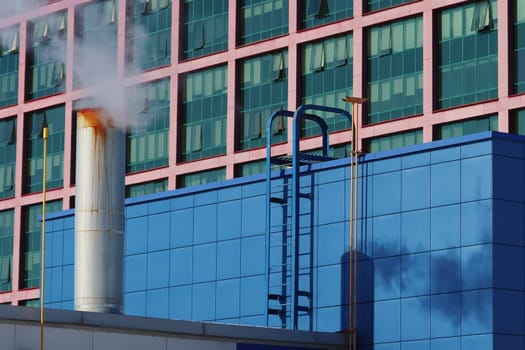 Factory chimney on the roof, against the background of a large building with windows.