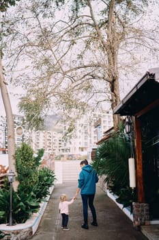 Mom and a little girl are standing in the park against the backdrop of high-rise buildings holding hands. Back view. High quality photo