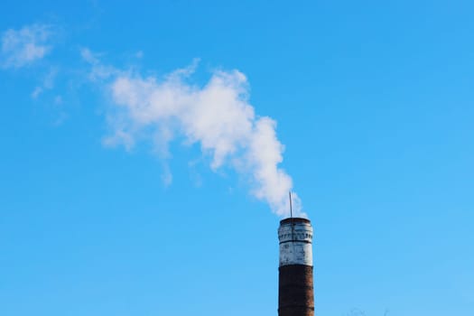 An old brick chimney with smoke against the background of a clear blue sky. Carbon Emissions Concept. Smoke from industrial chimney.