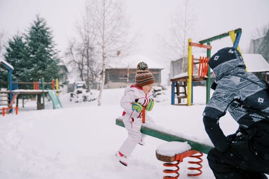 Mom and little girl ride on a snowy swing-balancer on a colorful playground. High quality photo