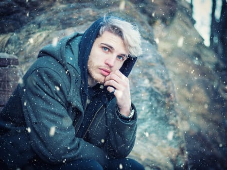 Portrait of young man in hoodie posing outdoor in winter setting with snow all around, looking at camera