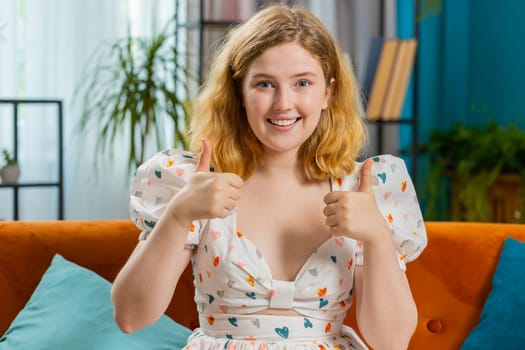 Like. Happy excited redhead woman looking approvingly at camera showing thumbs up, like sign positive something, good great news positive feedback. Young girl sitting on couch at home room. Horizontal