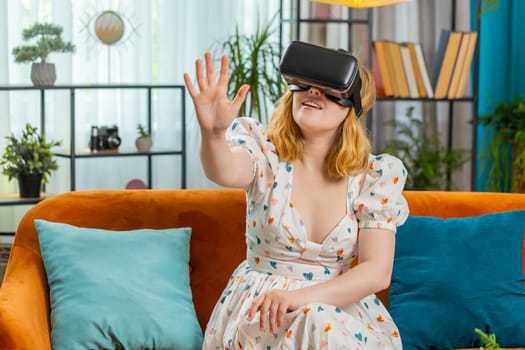 Woman using virtual reality futuristic technology VR app headset helmet to play simulation 3D 360 video game, watching film movie at modern home apartment. Girl in goggles sitting on couch. Horizontal