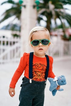 Little girl in sunglasses with a soft toy in her hand stands on the street. High quality photo