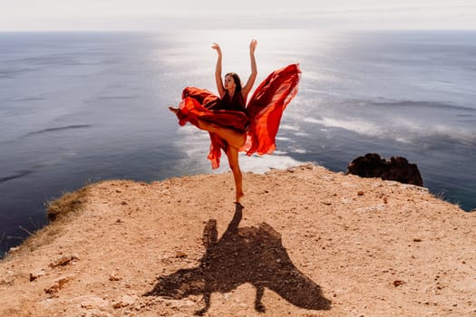 Woman red dress sea. Female dancer posing on a rocky outcrop high above the sea. Girl on the nature on blue sky background. Fashion photo