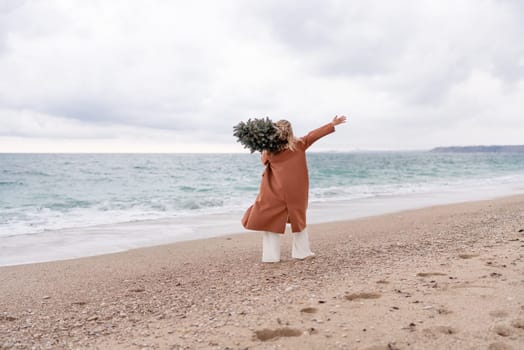 Blond woman Christmas sea. Christmas portrait of a happy woman walking along the beach and holding a Christmas tree on her shoulder. She is wearing a brown coat and a white suit