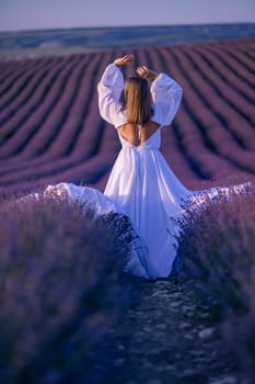 Woman poses in lavender field at sunset. Happy woman in white dress holds lavender bouquet. Aromatherapy concept, lavender oil, photo session in lavender.