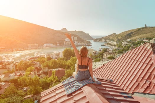 Woman sits on rooftop with outstretched arms, enjoys town view and sea mountains. Peaceful rooftop relaxation. Below her, there is a town with several boats visible in the water.