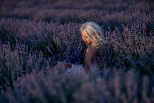 Blonde woman poses in lavender field at sunset. Happy woman in white dress holds lavender bouquet. Aromatherapy concept, lavender oil, photo session in lavender.