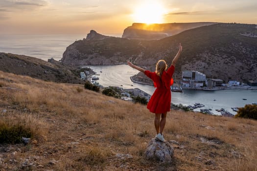 Happy woman standing with her back on the sunset in nature in summer with open hands posing with mountains on sunset, silhouette. Woman in the mountains red dress, eco friendly, summer landscape active rest.