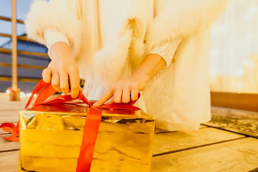 A woman in a white dress is holding a gold box with a red ribbon. She is wearing a crown on her head. The scene takes place in a room with a door and a window. The woman appears to be opening the gift box.