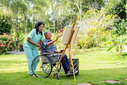 Wide shot of Asian nurse stand beside senior man sit on wheelchair and draw or paint for relax and revive good health of old people in outdoor with warm light.