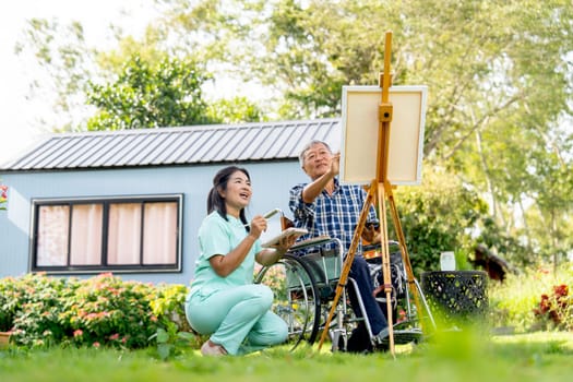 Asian nurse woman sit near senior man on wheelchair and cheer up him to paint the painting outdoor in the garden for relax and get good health.
