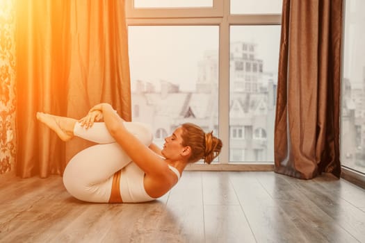 Young athletic attractive woman practicing yoga. Works out at home or in a yoga studio, sportswear, white pants and a full-length top indoors. Healthy lifestyle concept.