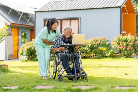 Wide shot of Asian nurse woman stand beside to take care senior man touch glassess and sit on wheelchair also stay to relax with laptop in the garden.