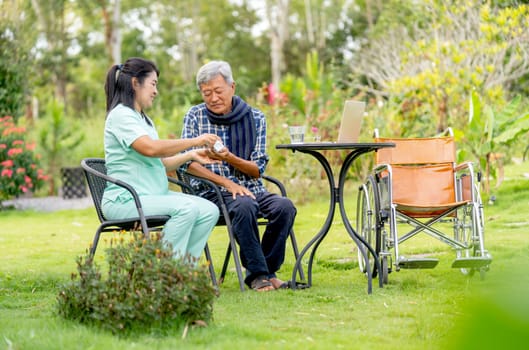 Asian nurse woman give the medicine pill from bottle to senior man and they sit on chair in the garden with wheelchair is set beside.