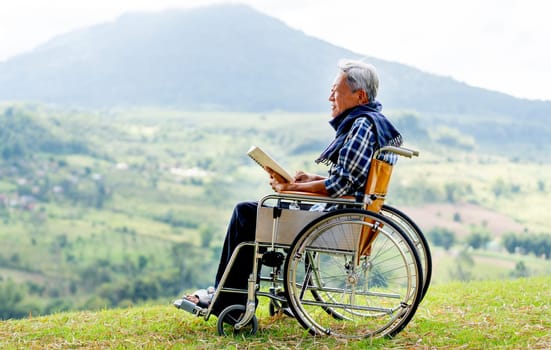 Asian senior man sit on wheelchair and read book also stay near cliff with meadow and mountain in the background with warm light and concept of nature health care.
