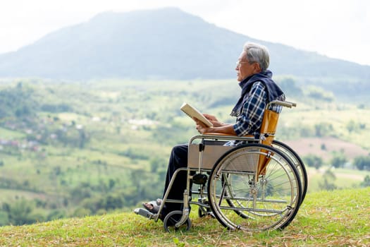 Asian senior man with eyeglasses look forward to relax after read the book and sit on wheelchair also stay near the cliff with grassfield and mountain as background.