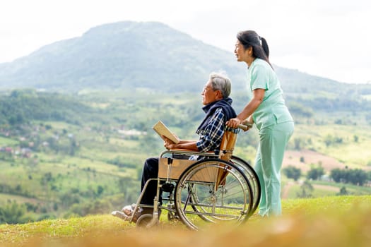 Asian nurse stand behide and hold wheelchair handle with senior man sit on wheelchair and both look forward and stay near cliff with village and mountain in the background.