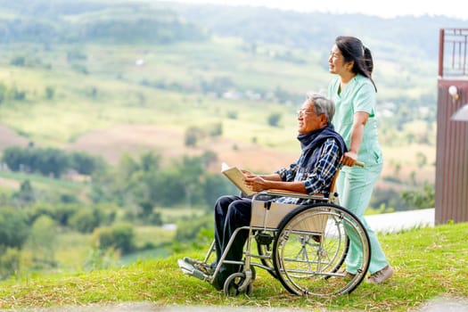 Asian nurse stand behide and hold wheelchair handle with senior man sit on wheelchair and both look forward and stay near cliff with village and mountain in the background.