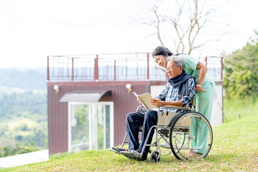 Asian nurse stand behide and look to the book hold by senior man on wheelchair and both look happy and stay near cliff with village and mountain in the background.