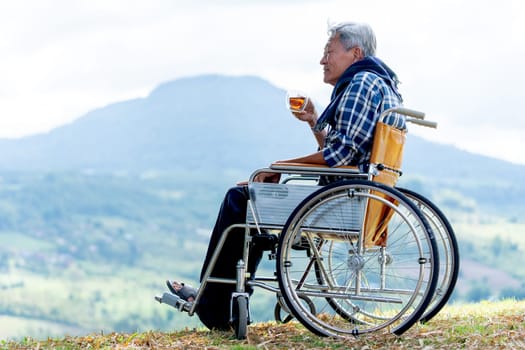 Asian senior man hold a cup of tea and sit on wheelchair with lood forward and stay near the cliff and mountain and grassfield as background.