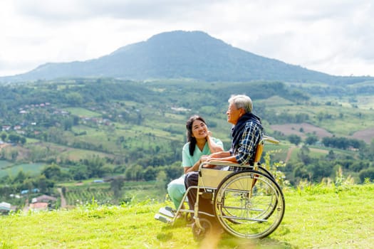 Asian nurse sit near senior man on wheelchair to take care for good health outdoor and both stay near the cliff with warm light.