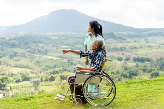 Asian nurse stand and look to the direction of senior man on wheelchair pointing and both stay near cliff with village and mountain in the background.