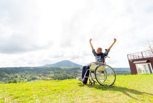 Wide shot of Asian senior man sit on wheelchair and action of raise his hands up like stretching also stay near cliff with mountain and grass field in the background.