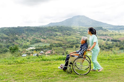 Asian nurse stand and hold wheelchair hand that sit by senior man and both stay near cliff with village and mountain in the background.