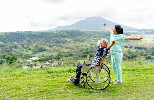 Side view of Asian nurse and senior man on wheelchair action of stretching together and both stay near the cliff with mountain and village as background.