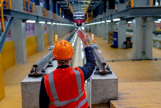 Back of technician worker with safety uniform show body signal to electrical or metro train and stand in front of railroad tracks in factory workplace.