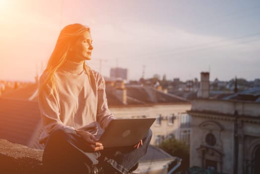 Woman freelancer uses laptop on cement wall outdoors against the sky and the roof of the city. The woman to be focused on her work or enjoying some leisure time while using her laptop