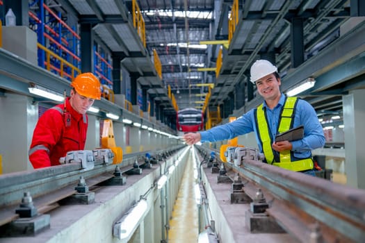 Professional engineer with smiling hold tablet and point to technician worker near railroad tracks of electrical or sky train in factory workplace.