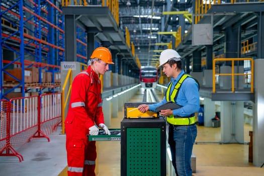 Technician and engineer worker discuss about tools and equipment with cabinet in front of railroad tracks of electrical or sky train in factory workplace.