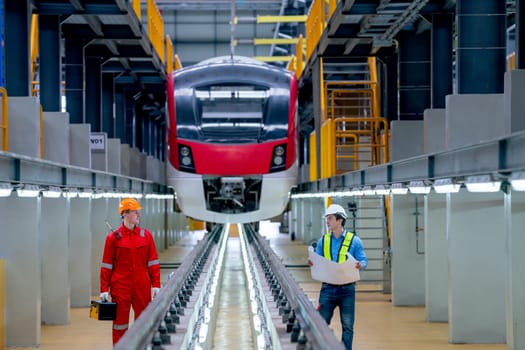 Professional engineer stand with hold plan or drawing document and discuss with technician worker near railroad tracks of electrical or sky train in factory workplace.