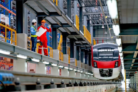 Wide shot of engineer hold drawing plan and technician worker stay and discuss beside railroad tracks of electrical or sky train in factory workplace.