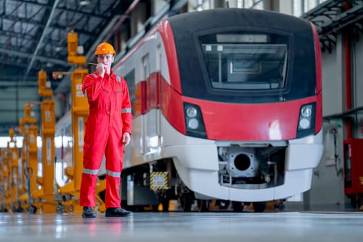 Professional technician worker stand in front of electrical or metro train and use walkie talkie to contact his team in train factory workplace.