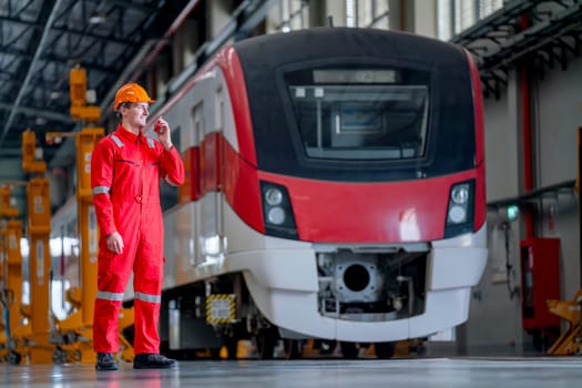 Professional technician worker stand in front of electrical or metro train and use walkie talkie to contact his team in train factory workplace.