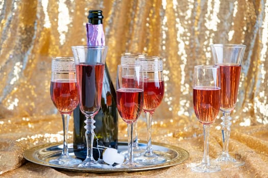 alcohol drinks concept. glasses of rose champagne and bottle on the tray on sparkling golden tablecloth