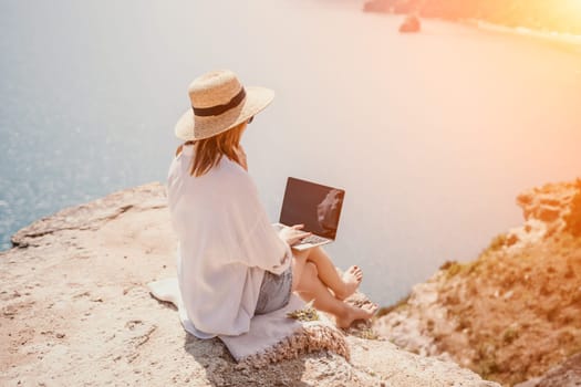 Successful business woman in yellow hat working on laptop by the sea. Pretty lady typing on computer at summer day outdoors. Freelance, travel and holidays concept.