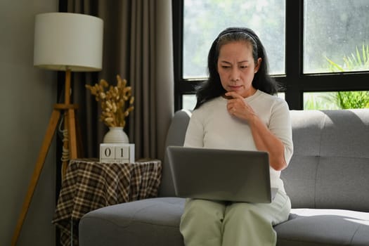 Happy middle age lady checking email or working on laptop while relaxing on the couch at home