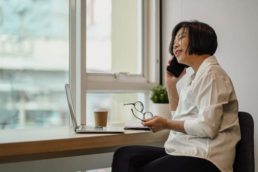 Smiling senior businesswoman holding glasses talking on mobile phone near office window.