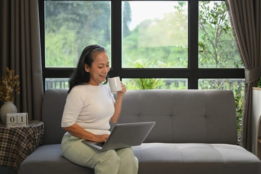 Smiling senior woman drinking hot tea and browsing internet on laptop at home