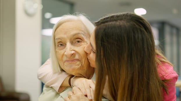 Smiling nurse that is a granddaughter kissing and embracing a senior woman in geriatrics