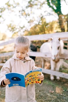 Pony looks from behind a fence at a little girl reading a book of fairy tales. High quality photo