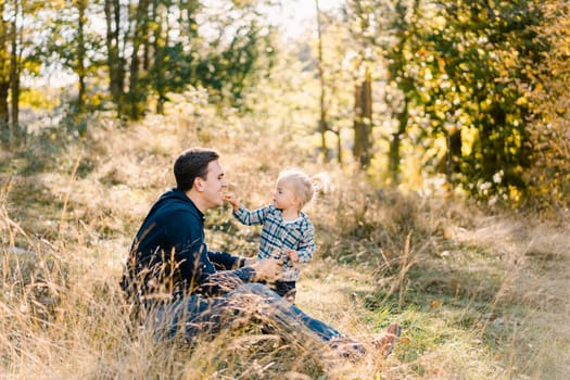 Little girl touches the face of her dad sitting on the lawn. Side view. High quality photo